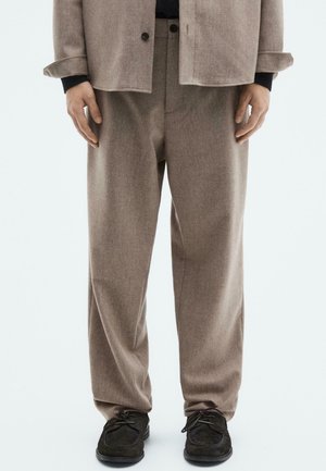 Person wearing loose taupe trousers, a matching jacket over a black top, and black lace-up shoes, standing against a plain background.