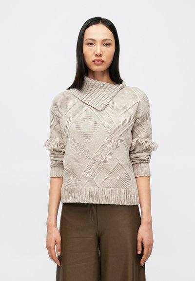 Femme aux longs cheveux bruns portant un pull en maille beige texturé avec des détails en franges sur les manches et un pantalon marron, debout devant un fond blanc.