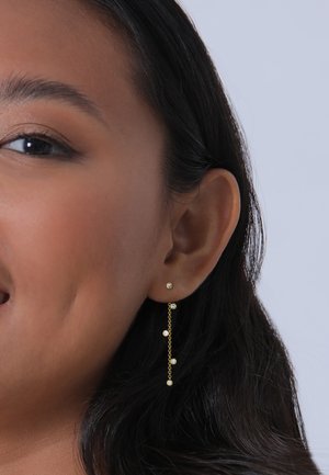 Goldene Tropfohrringe mit einer Kette, die fünf Perlen enthält, von denen zwei mit klaren Steinen verziert sind, ergänzen eine Nahaufnahme des Ohres.