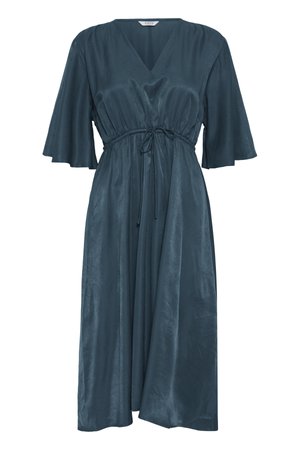 Femme aux cheveux foncés tirés en arrière porte des boucles d'oreilles en or, une robe turquoise à col en V avec manches volantes, un bracelet noir et une bague à la main gauche.