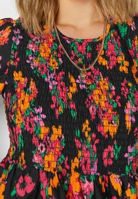 Donna che indossa un vestito floreale nero con motivi arancioni, rosa e verdi, e una collana in oro con un piccolo ciondolo rotondo.