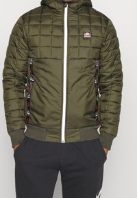 Man in een olijfgroene gewatteerde zip-up jas met logo-applicaties en zwarte joggers met witte logo's, met de handen in de zakken.