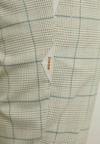 Beige och blått mönstrat tyg med en rutig design. Etiketten visar ”CHASIN’” i orange på en vit tagg. Texturerad, vävd struktur.