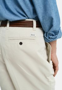 Pantalones de algodón beige con un cinturón de cuero trenzado marrón, que cuentan con un bolsillo trasero con botón y una etiqueta de GANT en la cintura.