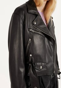 Zwart leren motorjack met zilveren ritsen, drukknoopsluitingen en riem, gedragen over een lichtpaarse top.