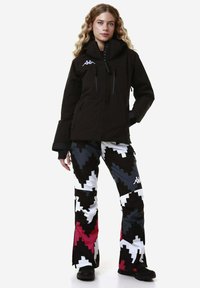 Giacca impermeabile nera con colletto alto, abbinata a pantaloni a fantasia con design zigzag in nero, bianco, grigio e rosa.