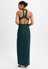 Donkergroene avondjurk met ingewikkelde kantdetails, een open rugontwerp en een aangesloten silhouet. Glad stof en een lengte tot de grond.