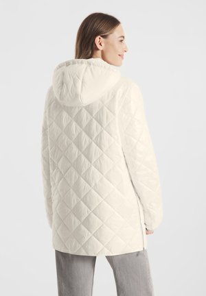 Kobieta w białej pikowanej kurtce z kapturem widziana od tyłu, w połączeniu z szarymi spodniami na jasnym, jednolitym tle.