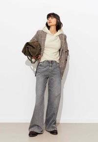 Blazer à carreaux marron sur un hoodie crème ; jeans évasés gris ; portant un sac vert foncé ; tissu texturé et coupe décontractée.