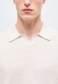 Visage inférieur et cou d'une personne portant un polo en maille blanche avec une petite ouverture en V au col.