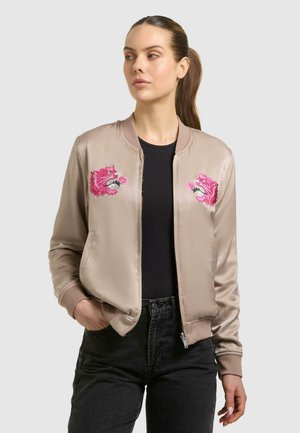 Frau mit langen braunen Haaren, trägt eine beigefarbene Satin-Bomberjacke mit Stickerei eines pinkfarbenen Tigerkopfs und Blumen, schwarzes Oberteil und schwarze Jeans.