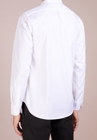 Camicia bianca a maniche lunghe con colletto button-down, orlo curvo e polsini con un bottone, realizzata in tessuto morbido, mostrata da dietro.