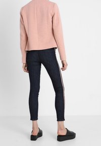 Veste texturée rose clair, jean skinny bleu foncé avec bandes latérales, et chaussures plates-formes noires sans lacets. Focus sur la vue arrière et la coupe.