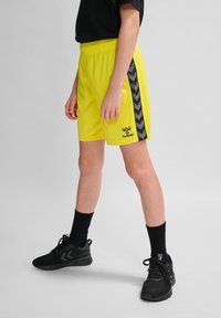 Neon-gelbe Sport Shorts mit schwarzen Chevron-Streifen an den Seiten, elastischem Bund und Logo auf der Vorderseite. Schwarze Sportschuhe und Socken.