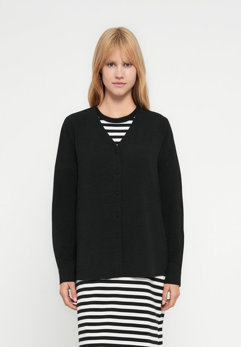 Cardigan noir à col en V et manches longues, en tissu texturé avec fermeture boutonnée à l'avant, porté sur une robe à rayures.
