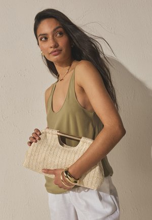 Kvinde med langt mørkt hår iført en grøn tanktop og hvide bukser, der holder en tekstureret beige clutch med guldarmbånd på håndleddet.