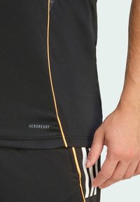 Svart atletisk t-shirt med en orange söm detalj på sidan, fuktavledande tyg, med "AEROREADY" logotyp. Hand vilar på en svart shorts med vita ränder.