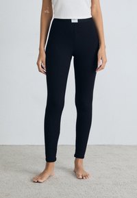 Black leggings with a stretchy fabric and a high waistband, featuring a logo tag at the front. They have a smooth, fitted design.