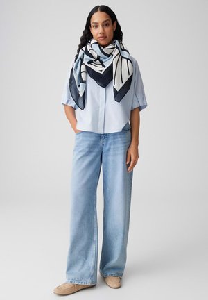 Femme portant une chemise bleu clair, un jean large en denim clair, des chaussures beiges et une grande écharpe à motifs bleu marine et blanc autour du cou.