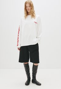 Hvid langærmet skjorte med rød tekst, parret med sorte knælange shorts og skinnende sorte støvler, hvilket skaber et moderne, afslappet look.