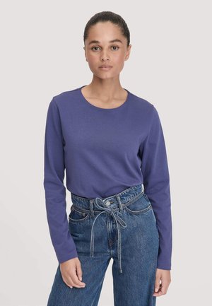 Lange mouw paarse top van een gladde stof, met een ronde halslijn. Gecombineerd met hoge blauwe jeans met een strik in de taille.