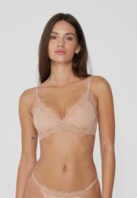 Femme aux longs cheveux bruns portant un bralette en dentelle nude et des sous-vêtements assortis sur un fond clair.