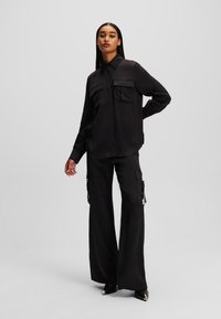 Schwarzes Hemd mit Knöpfen und zwei Vorderentaschen, kombiniert mit hochtaillierten Cargo-Hosen mit Seitentaschen und Bindebändern. Glattes, leichtes Material.