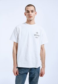 Bílé bavlněné tričko s krátkým rukávem, které má malé grafické provedení na levé části hrudi a volný střih. Na sobě má modré džíny.