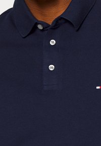 Camisa polo azul marino hecha de tela texturizada, con una tapeta de tres botones, mangas cortas y un pequeño logo bordado en rojo, blanco y azul.