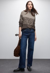 Grauer Strickpullover mit braunem Tierdruck, kombiniert mit dunkelblauen weiten Jeans. Das Model hält eine braune Wildledertasche und trägt schwarze Stiefeletten.