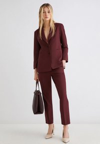 Donna che indossa un completo tailored bordeaux con giacca a due bottoni e pantaloni slim, tiene in mano una borsa bordeaux scuro e indossa décolleté beige.