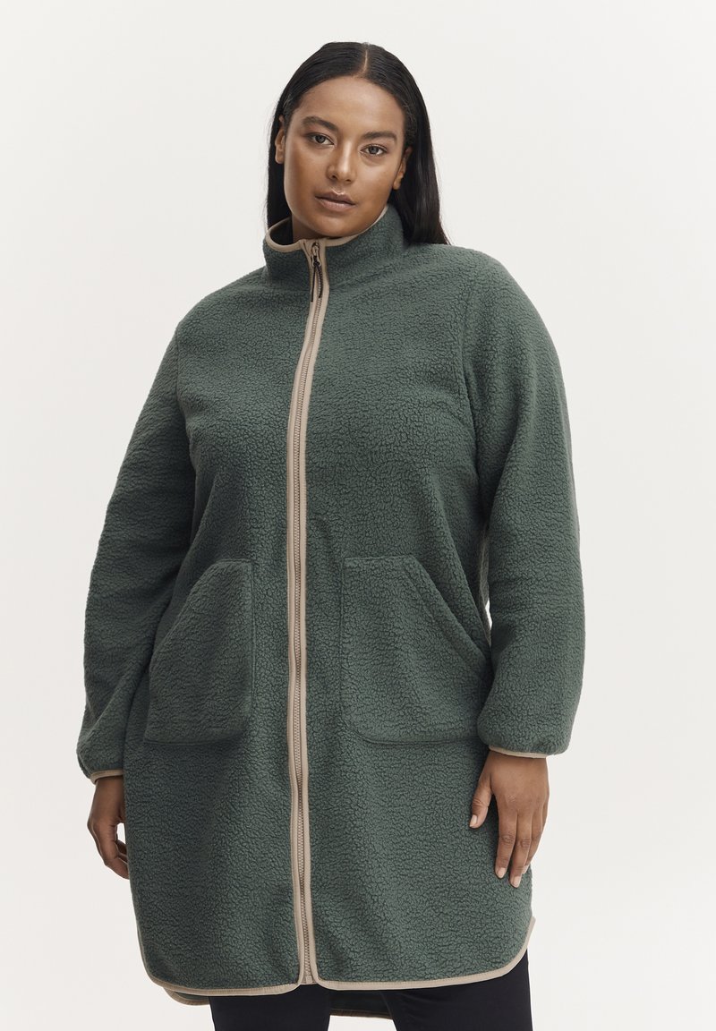 Abrigo de forro polar en verde oscuro con un cuello alto, cremallera beige de contraste, dos bolsillos delanteros y tela texturizada. Mangas largas y largo hasta la rodilla.