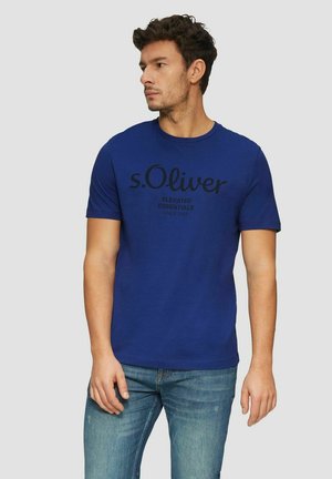 Blue cotton t-shirt with crew neckline, featuring a black printed logo text design. Styled with light blue jeans, model stands against a plain background.