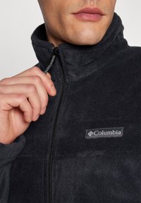 Veste en polaire noire avec un col montant, fermeture éclair sur le devant et patch logo Columbia. Texture douce avec une finition lisse.
