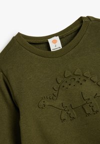 Olivgrön sweatshirt med en texturerad dinosauriedesign. Ribbad rund halsringning och en etikett märkt "respect life" synlig inuti.