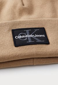 Braune Strickmütze mit strukturiertem Stoff. Sie verfügt über ein schwarzes rechteckiges Patch mit dem Logo "Calvin Klein Jeans" in weißer und grauer Schrift.