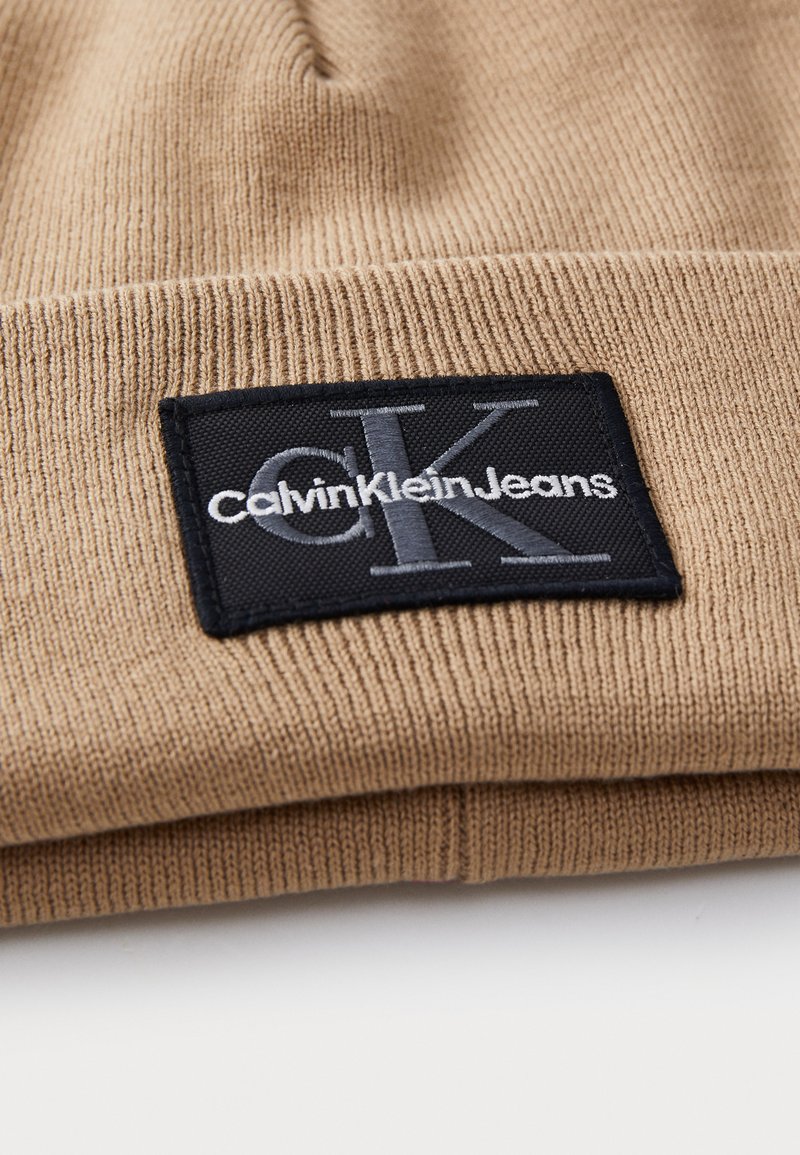 Brązowa dzianinowa czapka z teksturowanym materiałem. Posiada czarną prostokątną naszywkę z logo "Calvin Klein Jeans" w białym i szarym kolorze.
