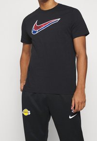 Camiseta negra de Nike con un logo de swoosh colorido y estampado; combinada con pantalones deportivos negros que presentan un logo de los Lakers. Textura suave, diseño de cuello redondo.