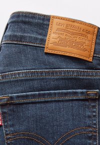 Jeans de mezclilla azul oscuro con un parche de cuero marrón con texto y diseños en relieve, costuras visibles y una etiqueta roja en el lateral.