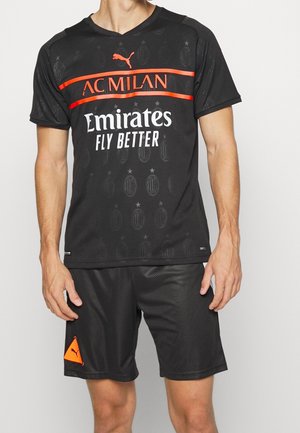 Osoba ubrana w czarną koszulkę piłkarską AC Milan oraz pasujące spodenki z pomarańczowymi logo Puma.