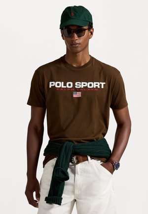 Bruine katoenen t-shirt met "POLO SPORT" graphic in wit en rood, witte shorts, groen vest om de taille, groene pet, donkere zonnebril.