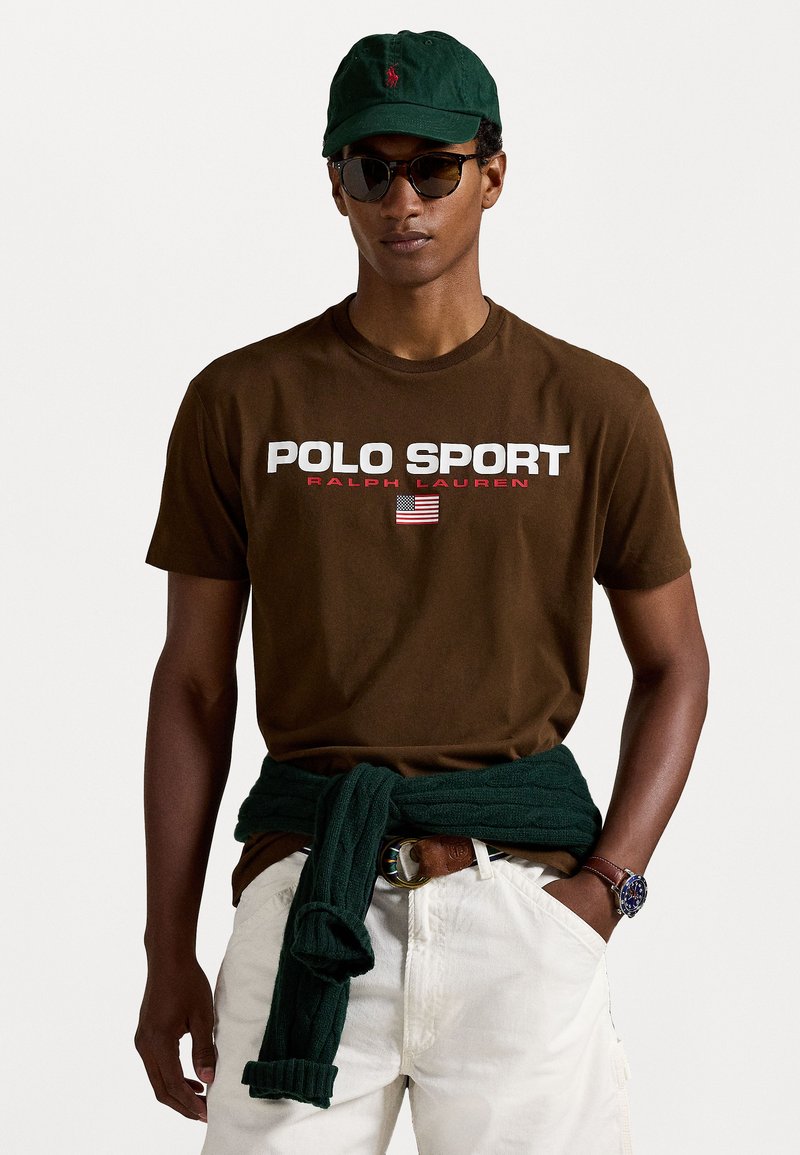 Camiseta de algodón marrón con gráfico "POLO SPORT" en blanco y rojo, pantalones cortos blancos, suéter verde atado a la cintura, gorra verde, gafas de sol oscuras.