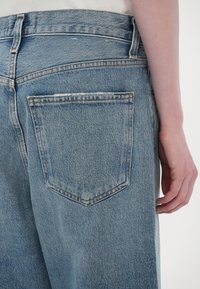 Jean en denim bleu clair vu de dos avec une poche arrière visible et la main droite d'une personne reposant à côté de la hanche.