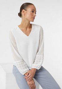 Biała bluzka z dekoltem w serek i długimi przezroczystymi rękawami z wzorem w kratkę na mankietach, noszona z jasnoszarymi spodniami. Gładka tekstura.