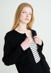 Čierna bunda z vlnenej zmesi s okrúhlym výstrihom, otvoreným predným dielom a dvoma prednými vreckami, vrstvená nad bielym pruhovaným topom. Hladká textura.
