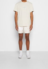 Beige t-paita, jossa on sininen painatus, yhdistettynä vaaleanbeigeihin shortseihin, joiden sivussa on logo. Yhdistettynä valkoisiin korkeatoppiin lenkkarikenkiin.