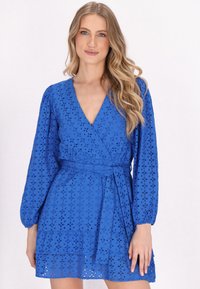 Sininen kääntömekko, jossa on pitkät hihat, silmukkaleikkauksia, syvä V-pääntie ja vyötetty waist. Kerroksellinen röyhelöhelma.