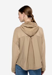 Sudadera con capucha beige que presenta un corte holgado, textura suave y un panel de tela contrastante en la parte trasera, combinada con pantalones de mezclilla azul.