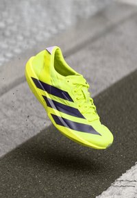 Φωτεινό κίτρινο αθλητικό παπούτσι με μωβ ρίγες, ελαφρύ σχέδιο, υφή στο επάνω μέρος και εμφανές σύστημα κορδονιών. Κρεμασμένο πάνω από μια επιφάνεια.