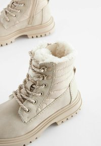 Beige enkellaarsjes van textiel met nepbontvoering, dikke veters en een robuuste zool. Beschikt over een gevoerd kraag en stikseldetails.